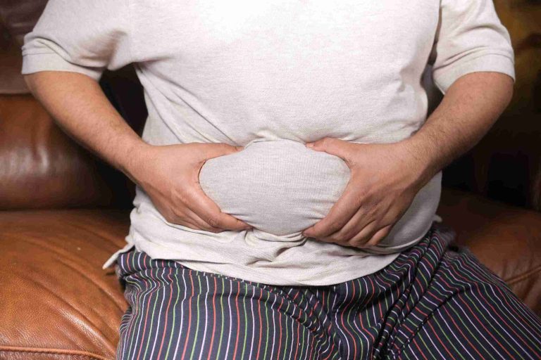 Male sitting on a couch and holding his bloated stomach.
